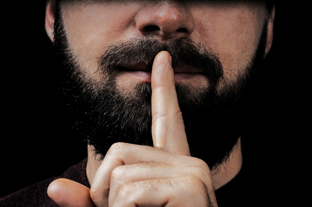 Bărbat cu barbă care face un semn cu degetul, indicând faptul că păstrează un secret