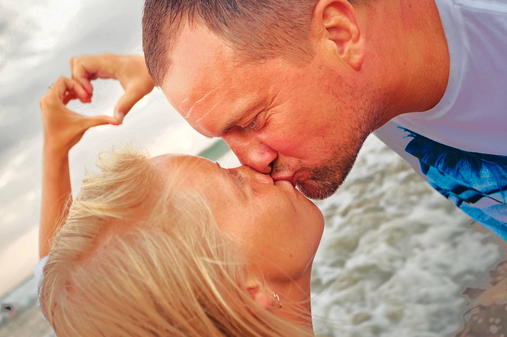 Parteneri care se sărută pe plajă, în timp ce formează o inimă din apropierea mâinilor