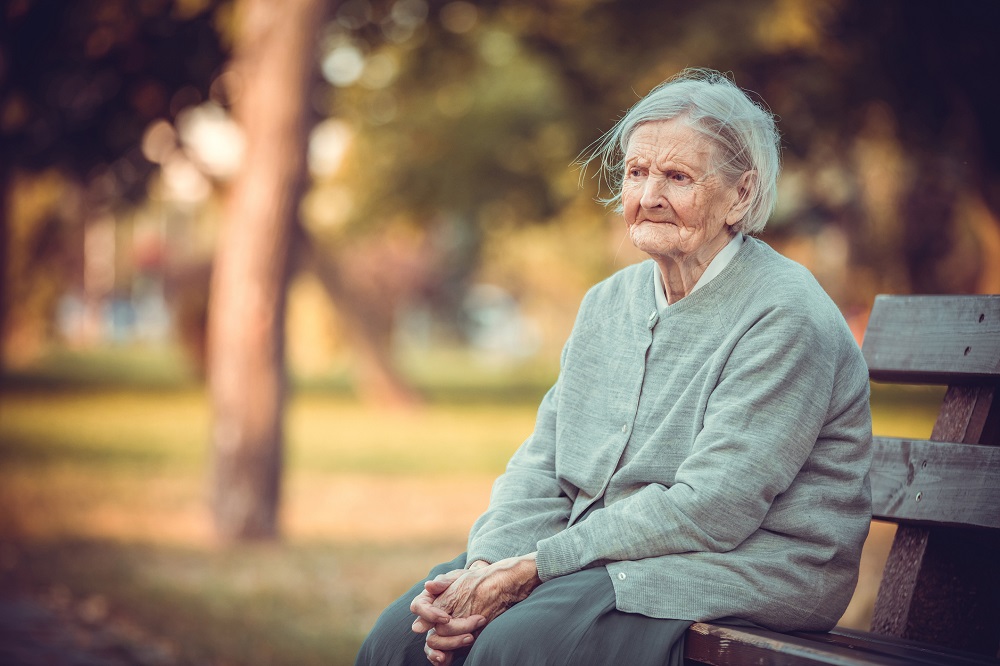 Femeie în vârstă și tristă care stă pe o bancă în parc și își ține palmele încrucișate.