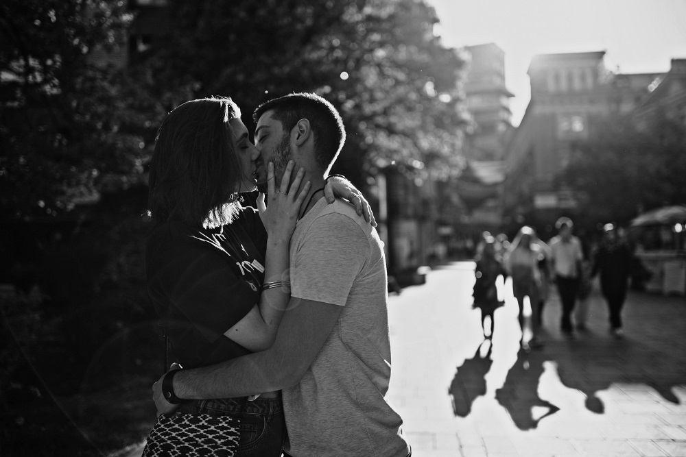 Tineri îndrăgostiți care se sărută pe stradă, în timp ce alți oameni trec pe lângă ei