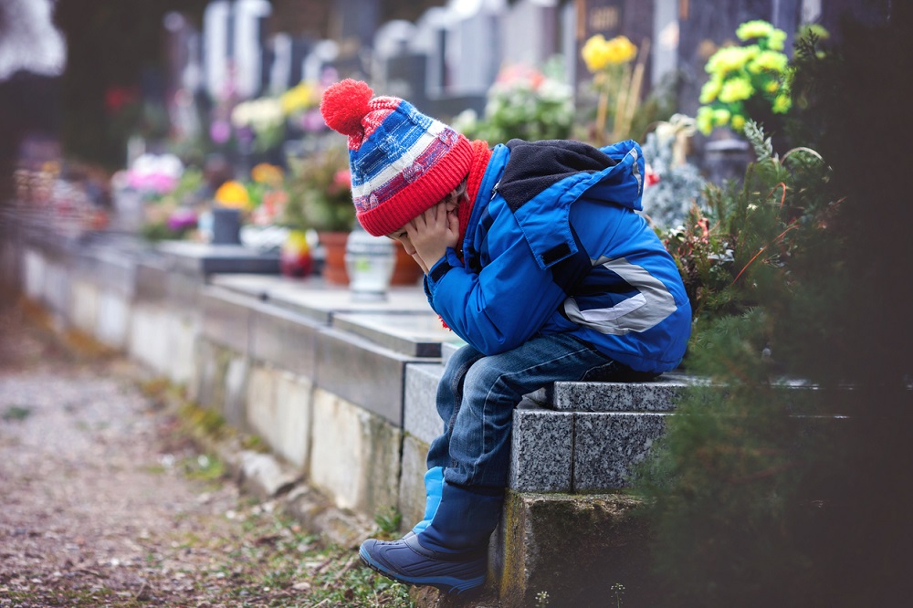 Copil trist, stând în cimitir, lângă mormântul mamei sale
