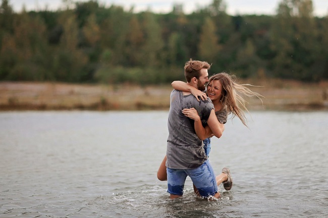 Parteneri îndrăgostiți care se distrează și râd, stând în apă, în timp ce bărbatul cară femeia în spate