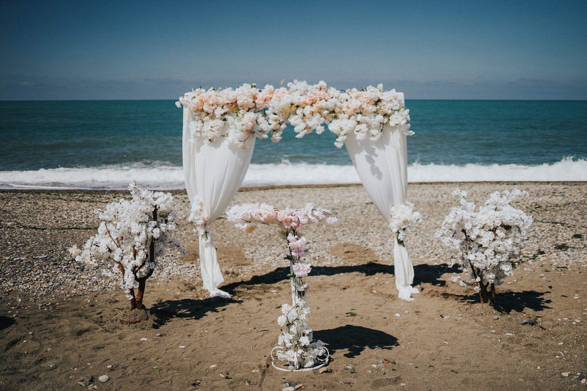 Altar de nuntă montat pe plajă, decorat cu flori albe și copăcei înfloriți