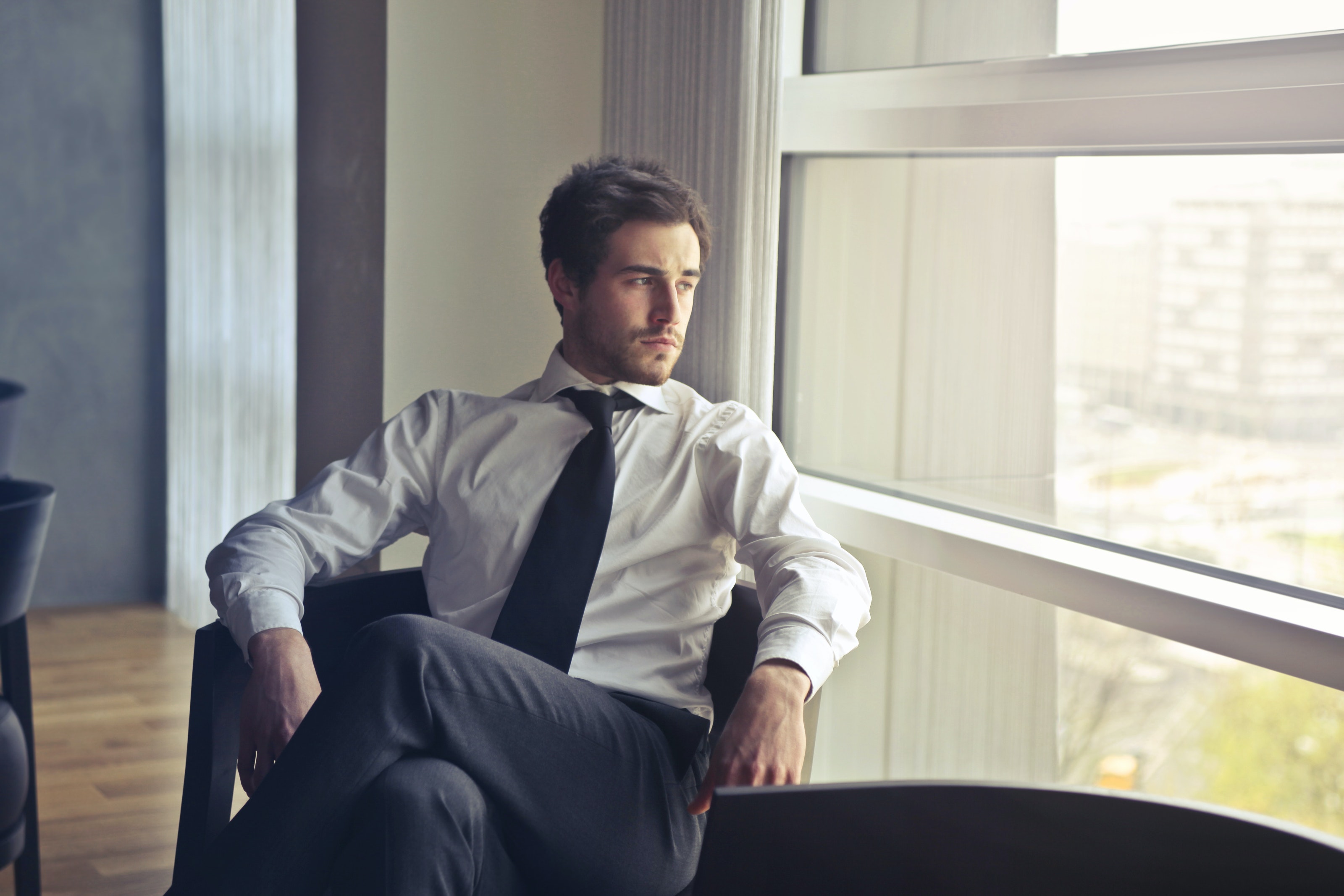 Bărbat îmbrăcat în costum, stând pe un scaun în birou și privind pe fereastră.