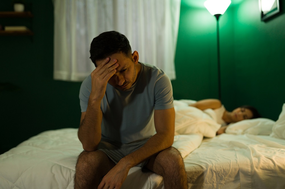 Bărbat supărat care stă pe marginea patului și se gândește la probleme, în timp ce partenera lui doarme.