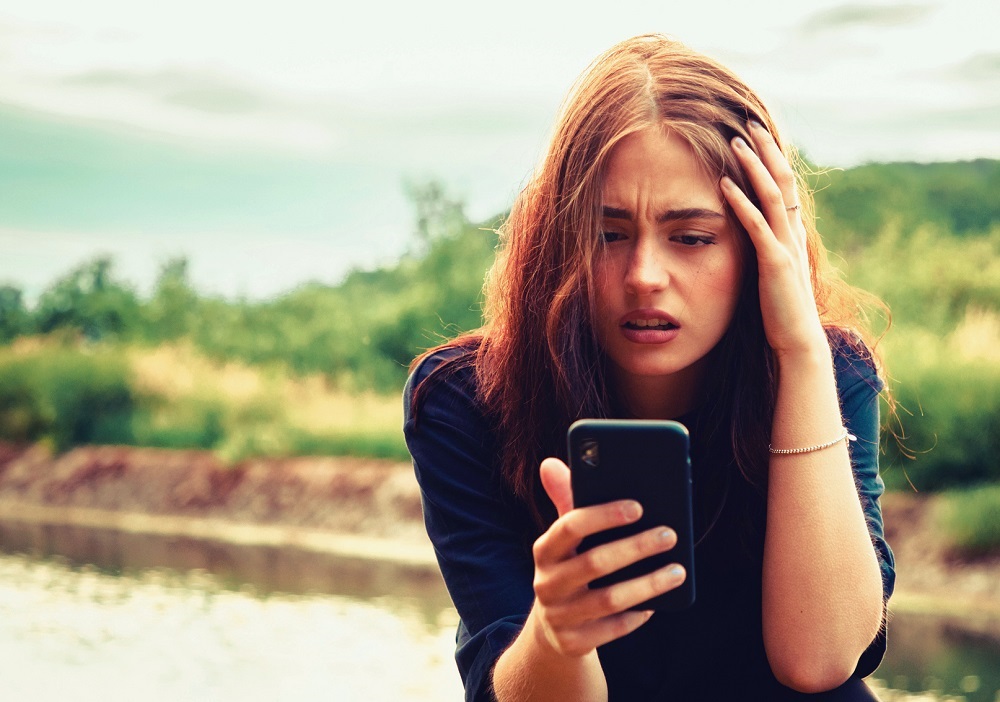 Femeie blondă care se uită confuză și cu disperare spre telefonul mobil, în timp ce își pune o mână în cap.