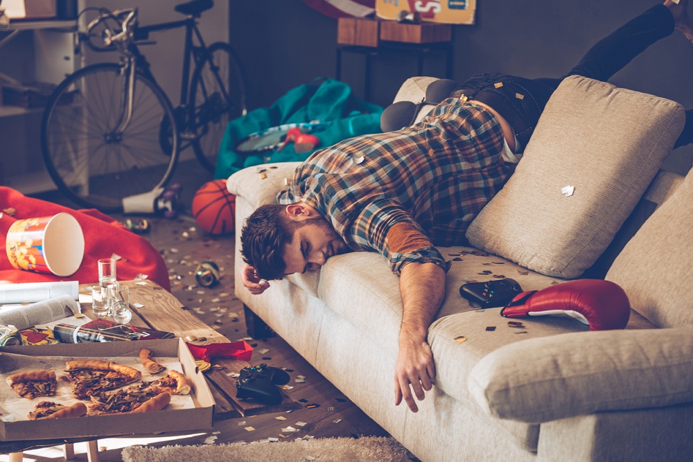 Bărbat care doarme pe canapea într-o cameră dezordonată și plină de mizerie, cu resturi de mâncare și ambalaje aruncate peste tot.