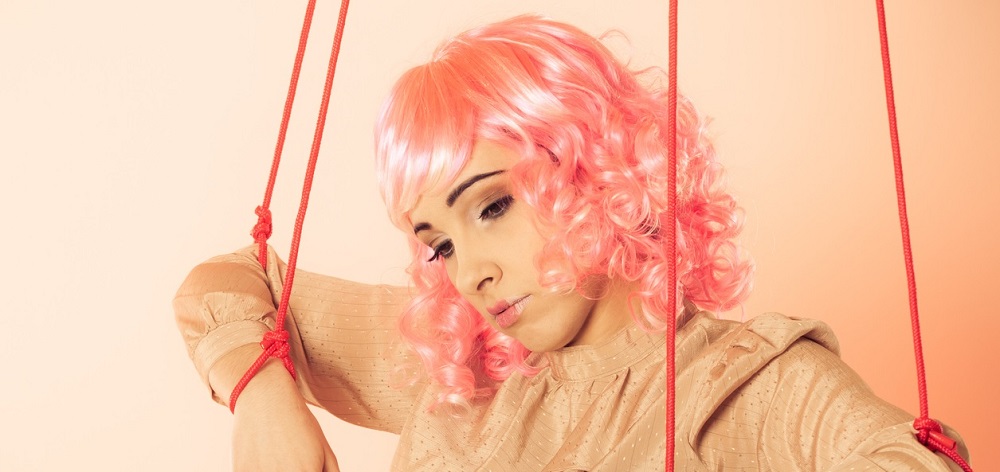 Femeie cu perucă roz, lăsându-se manevrată ca o marionetă, încercând să ilustreze conceptul de manipulare.