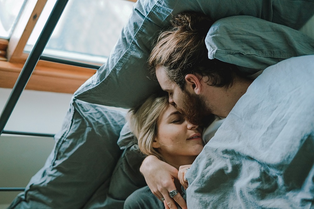 Cuplu care stă îmbrățișat în pat, în timp ce bărbatul își sărută partenera pe frunte.