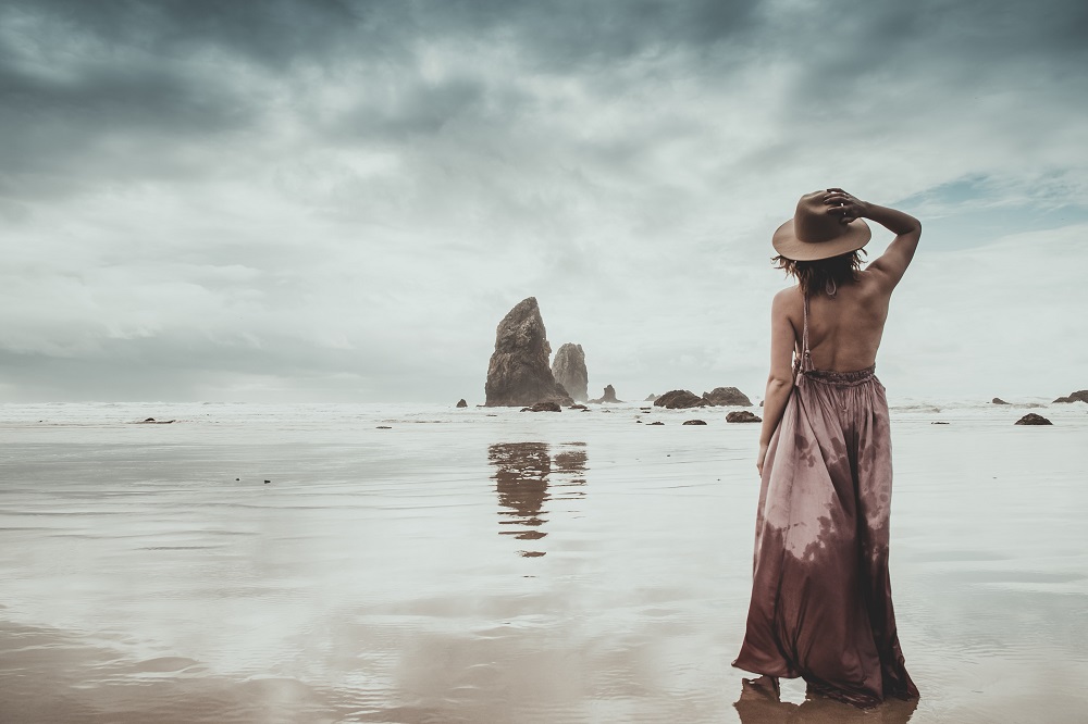 Femeie singură pe plajă, stând cu spatele și admirând peisajul.