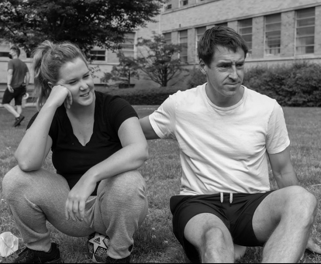 Imagine alb-negru cu Amy Schumer și Chris Fischer stând pe iarbă.