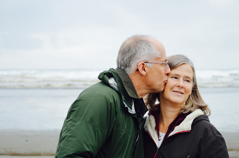 Bărbat în vârstă care își sărută partenera pe obraz, în timp ce stau pe malul mării