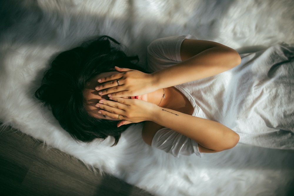 Femeie care stă întinsă în pat și își acoperă fața cu mâinile.