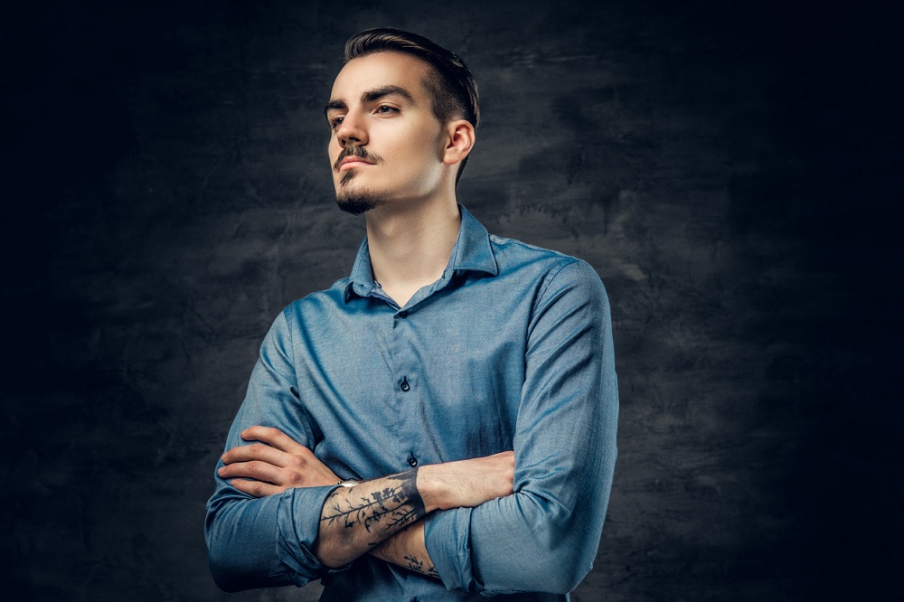 Portret al unui bărbat serios, cu tatuaje, care stă cu brațele încrucișate și poartă o cămașă albastră.