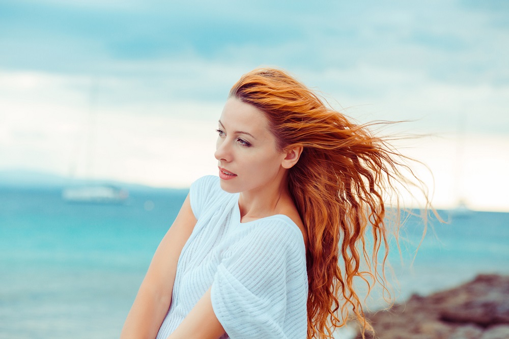 Imagine din profil cu o femeie roșcată, cu păr lung și ondulat lejer, în timp ce privește în zare de pe malul mării.