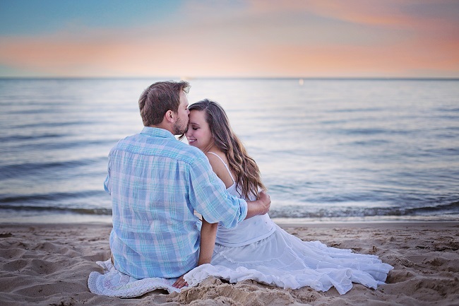Bărbat care își sărută femeia pe frunte, în timp ce stau îmbrățișați pe malul mării.