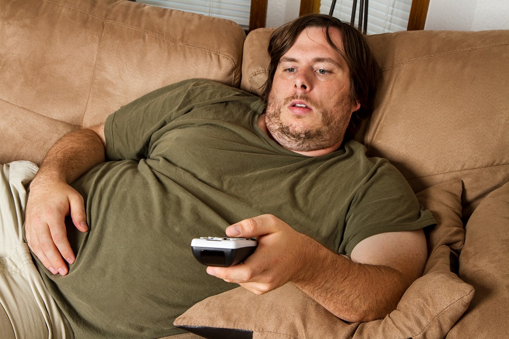 Bărbat supraponderal care lenevește pe canapea și se uită la televizor.