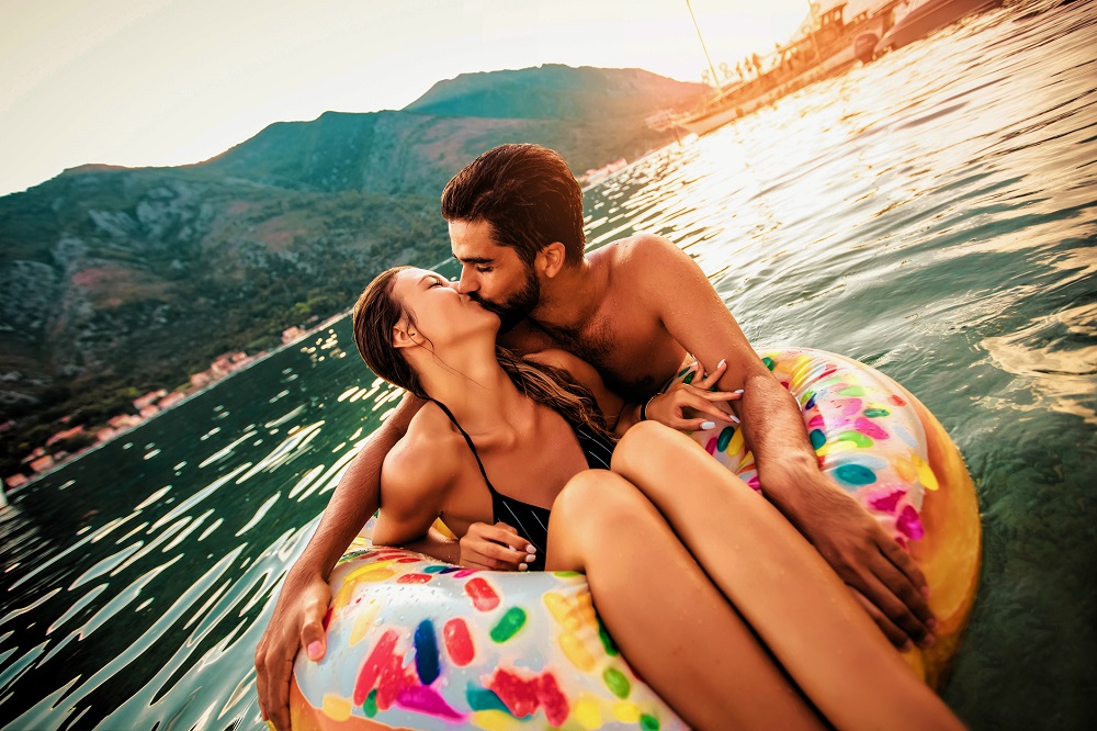 Îndrăgostiți care se sărută în mare, în timp ce femeia plutește pe un colac multicolor.