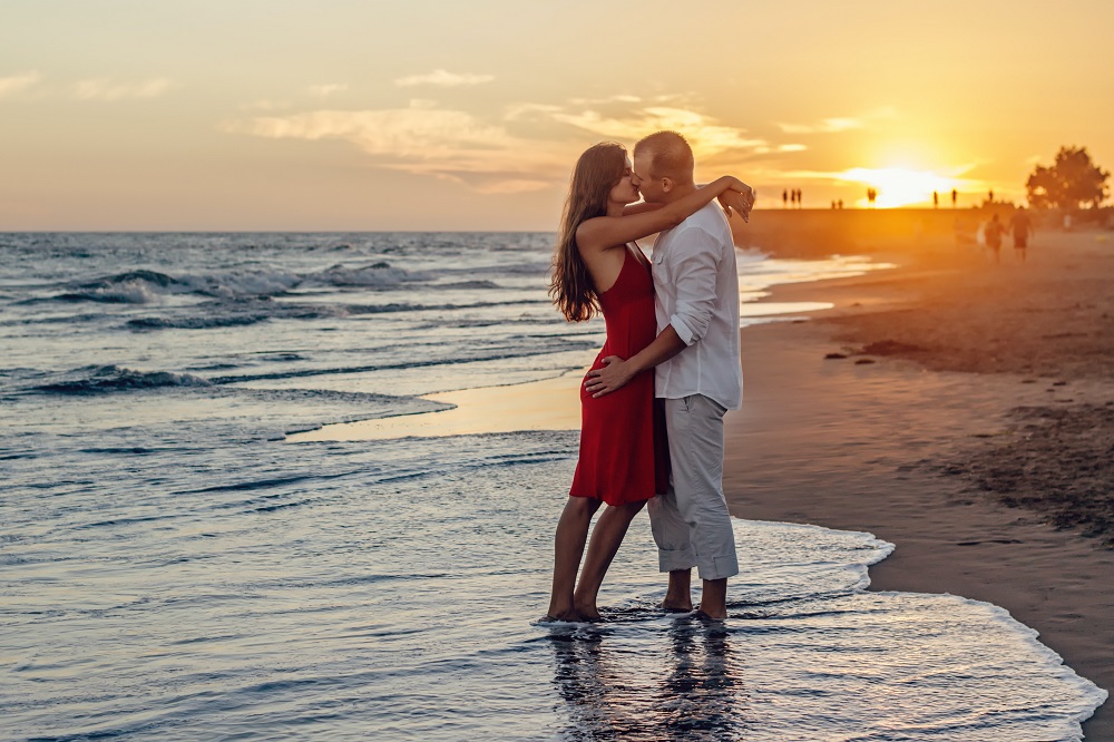 Îndrăgostiți care se sărută pe plajă, în timp ce stau cu picioarele în mare.