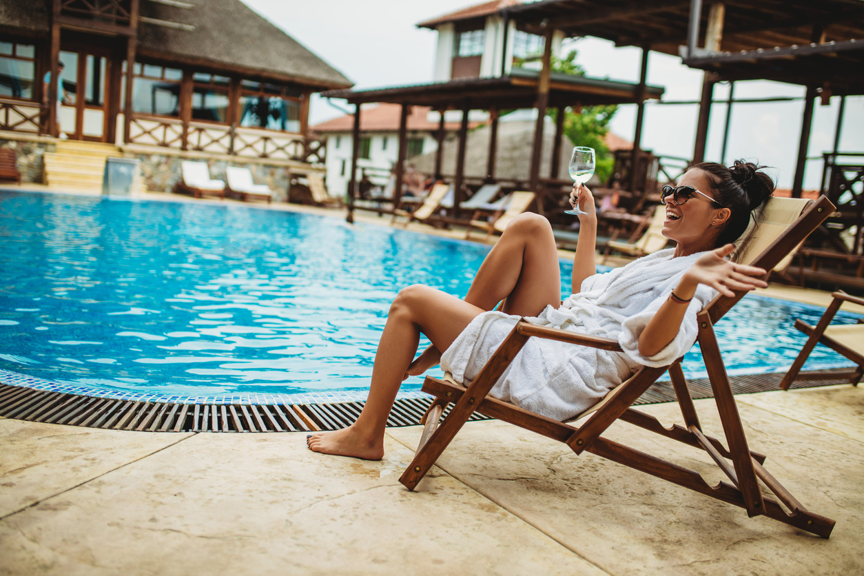 Femeie fericită care stă la piscină îmbrăcată într-un halat alb și cu un pahar de vin alb în mână