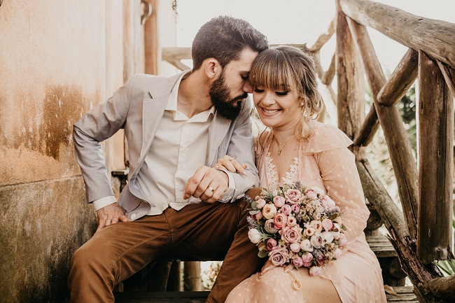 Moment tandru între îndrăgostiți în ziua nunții, bărbatul își sprijină fruntea pe tâmpla femeii, iar ea zâmbește și ține un buchet de flori în mână.