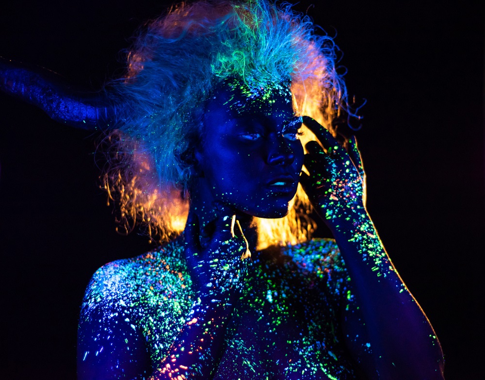 Femeie machiată pe chip și pe trup în culori fluorescente, care poartă lentile de contact albastre și perucă.