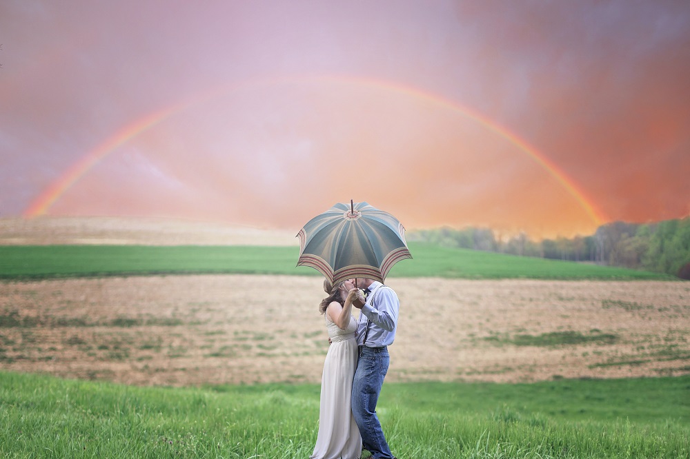 Parteneri care se sărută sub o umbrelă, după ploaie, cu un curcubeu în fundal.