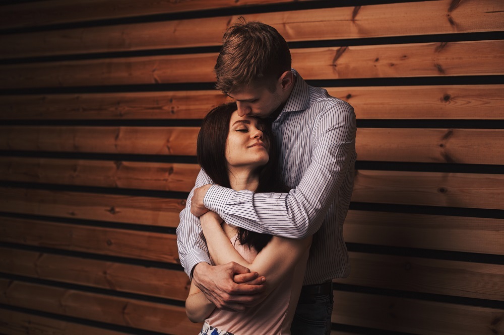 Bărbat care își strânge partenera în brațe și o sărută pe frunte, în timp ce stă lângă un perete maro.