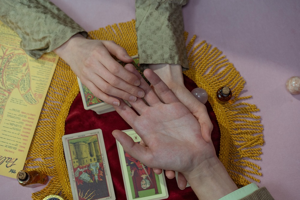 Femeie care îi citește în palmă unui bărbat, în timp ce mâinile lor sunt întinse peste o masă pe care sunt așezate cărți de tarot.
