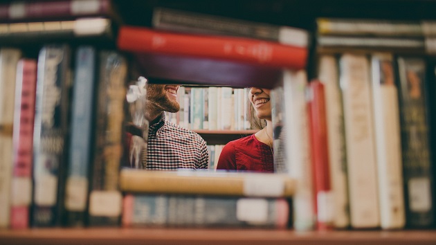 Parteneri care se privesc în ochi și zâmbesc, în timp ce se plimbă printre rafturile unei biblioteci.