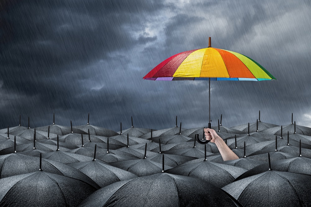 Persoană care se diferențiază de mulțime prin intermediul unei umbrele colorate. Imagine care ilustrează optimismul într-o mare de pesimism.