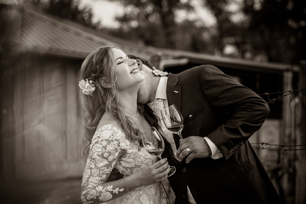 Bărbat care își sărută soția pe gât în ziua nunții, în timp ce țin în mână paharele cu șampanie.