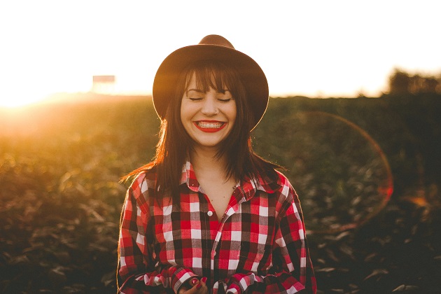 Femeie fericită, cu un zâmbet larg, purtând o cămașă colorată în carouri și o pălărie de culoare închisă