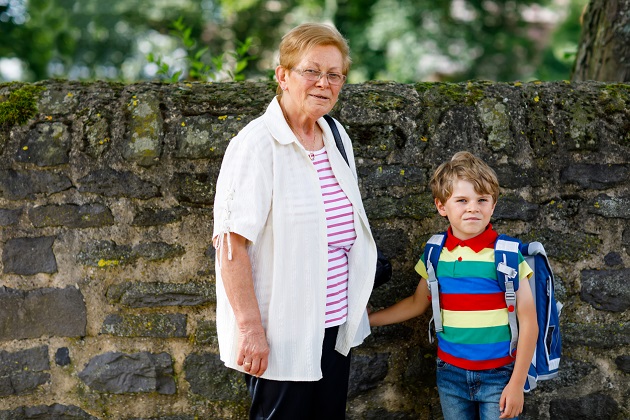 Băiețel cu ghiozdanul în spate care o ține de mână pe bunica lui.