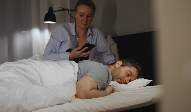 Femeie care se uită noaptea în telefonul partenerului, în timp ce el doarme.
