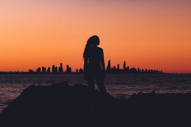 Silueta unei femei care stă pe plajă și admiră orașul care se vede în depărtare.