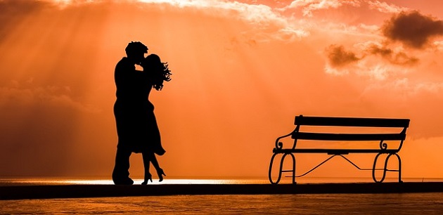 Siluetele unor îndrăgostiți care se sărută în razele soarelui, lângă o bancă.