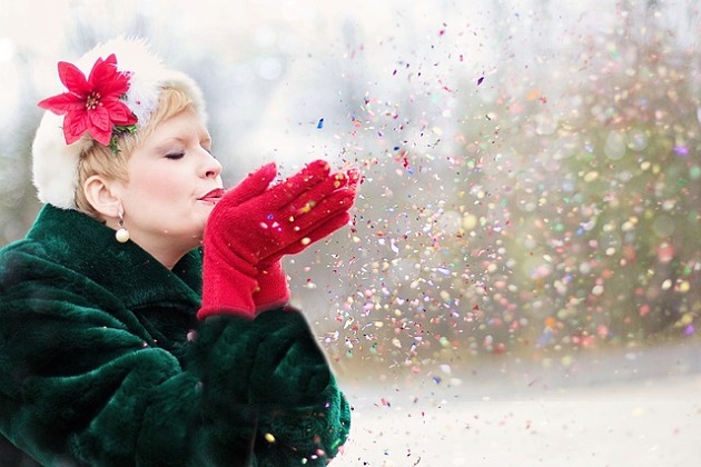 Femeie blondă care suflă în confetti colorate, în timp ce poartă mănuși roșii și căciulă albă care are atașată o crăciuniță într-o parte