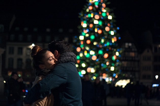Îndrăgostiți care se îmbrățișează în fața unui brad de Crăciun împodobit cu luminițe colorate și amplasat afară