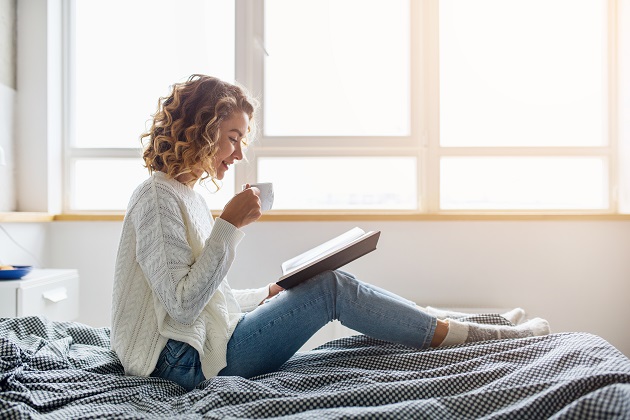 Femeie blondă și creață care citește o carte și bea cafea, în timp ce stă în pat.