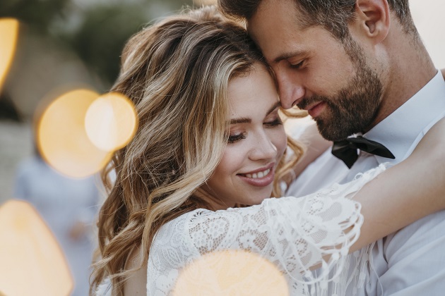 Poză de cuplu la nuntă, în care mireasa se prinde de gâtul mirelui și zâmbește, în timp ce el își sprijină fruntea de tâmpla ei.
