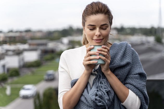 Femeie care stă pe balcon și își savurează cafeaua la aer curat, în timp ce ține o pătură gri pe umăr.