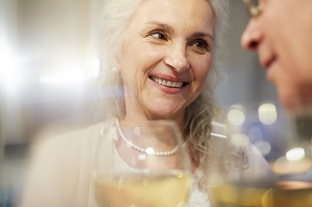 Femeie în vârstă zâmbitoare, îmbrăcată în alb și cu perle albe la gât, care ciocnește un pahar de șampanie.