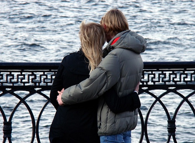 Parteneri care stau îmbrățișați pe un pod și admiră valurile agitate din fața lor.