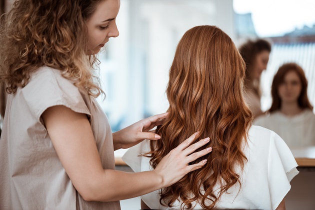 Femeie cu părul castaniu care stă pe scaun la salon, în timp ce hairsylista o coafează