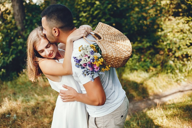 Bărbat care își îmbrățișează partenera și o sărută pe tâmplă, în timpul unei plimbări prin pădure, după ce ea a cules un coșuleț cu flori.
