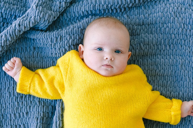 Bebeluș îmbrăcat în galben, așezat pe patul acoperit cu o pătură albastră