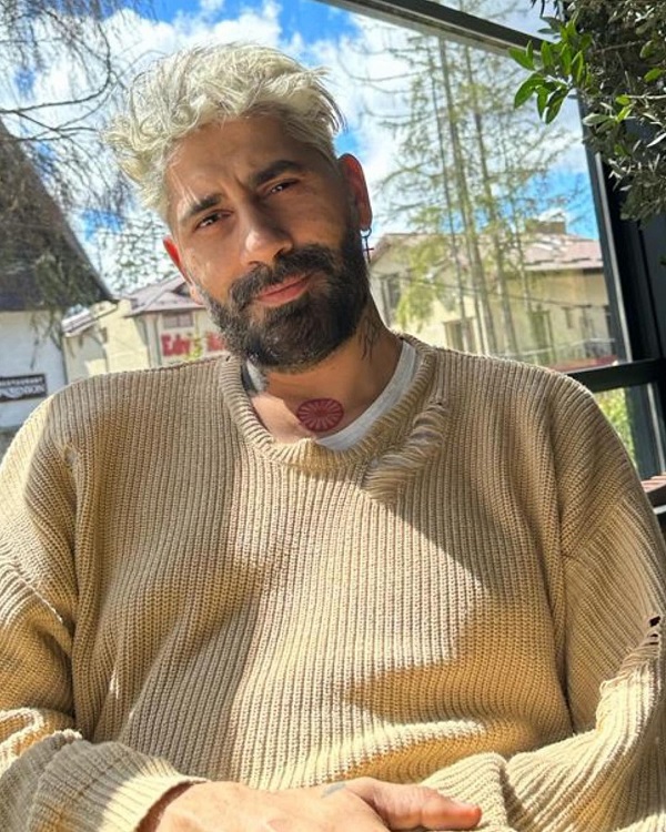 Connect-R stând la o terasă, îmbrăcat într-un pulover bej care se asortează cu părul său blond