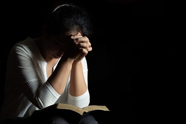 Femeie care se roagă cu palmele încrucișate, în timp ce are Biblia așezată pe genunchi
