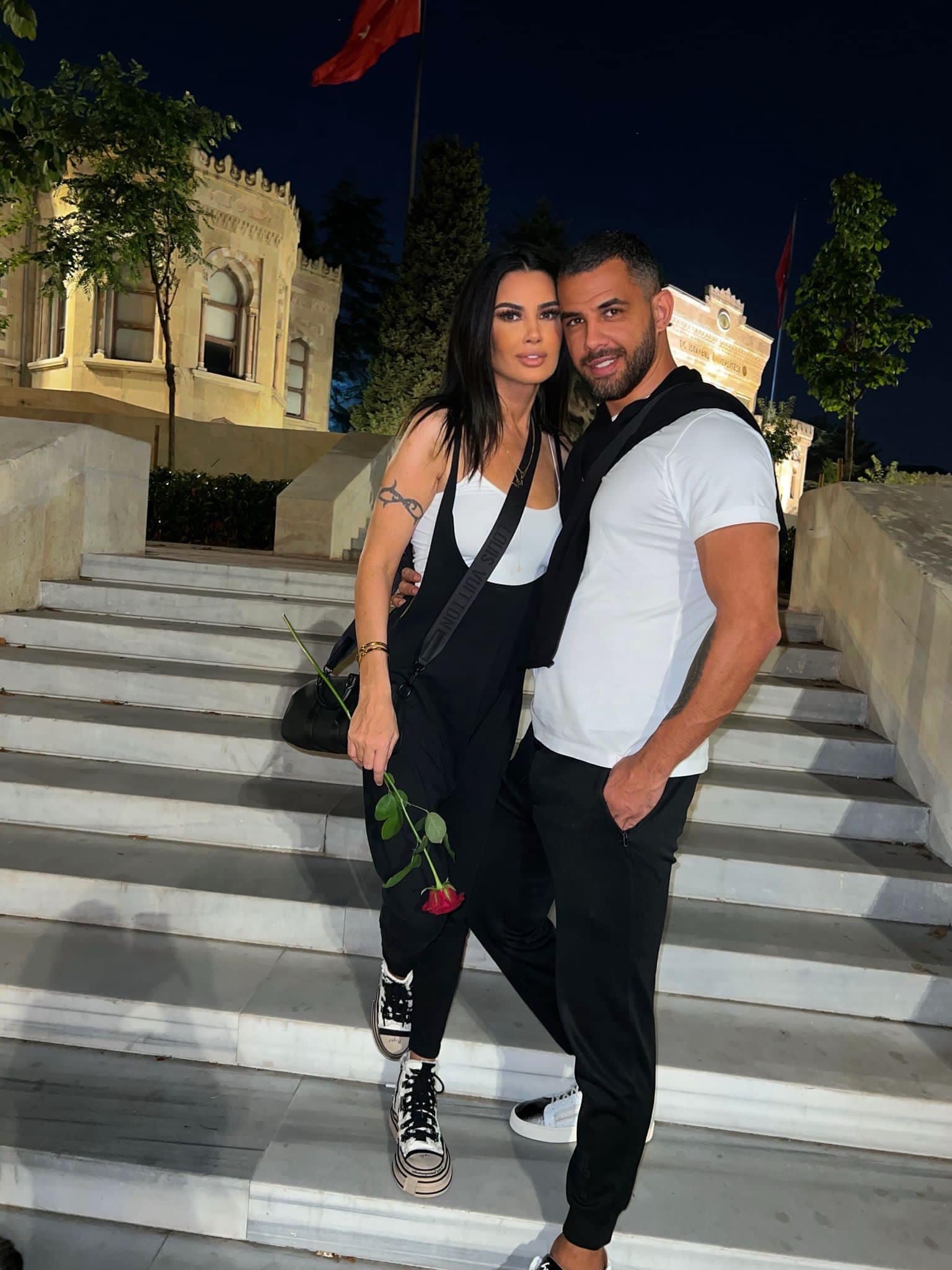 Oana Zăvoranu cu un trandafir roșu în mână, ținută în brațe de Alex Ashraf pe niște trepte.
